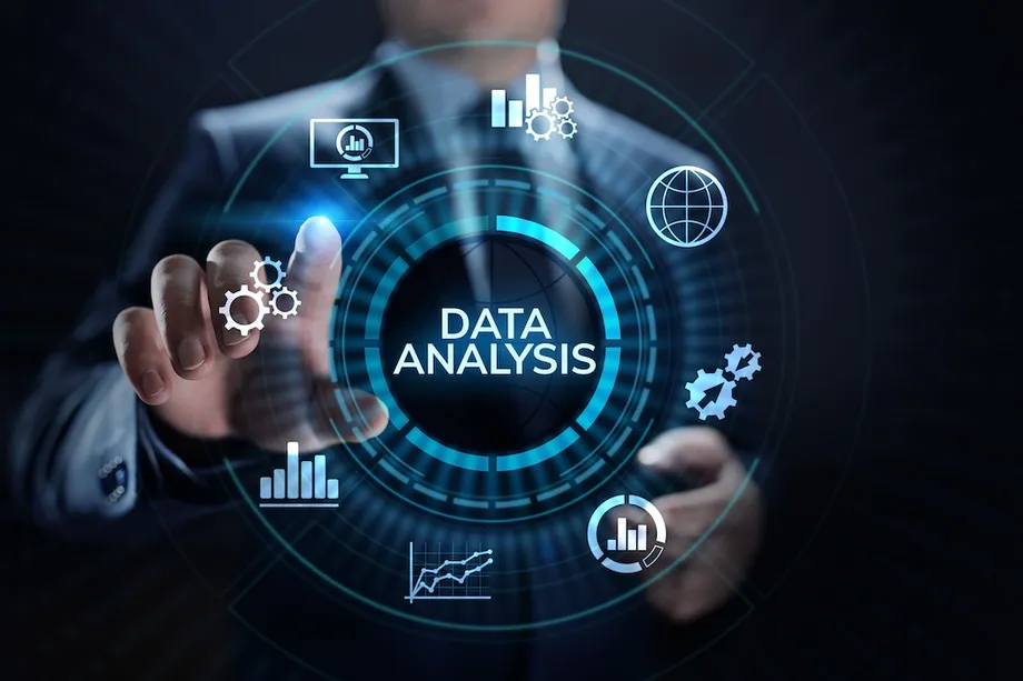 A businessman pointing at a glowing "Data Analysis" holographic interface surrounded by icons for charts, gears, a globe, and analytics dashboards, representing business intelligence and data-driven decision making.